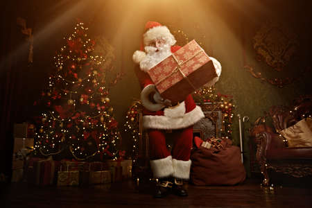 Santa Claus brought gifts for Christmas. He stands in a night room with beautiful Christmas tree and fireplace. Christmas and New Year concept.の写真素材