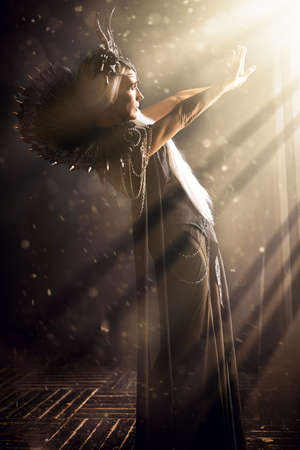 Fantasy World. A beautiful majestic black queen, a witch in a rich headdress and a rich black dress, stands in the rays of light in a dark castle.の写真素材