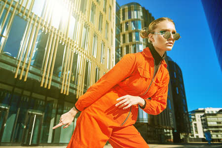 Bright youth style. Modern fashionable girl in bright orange jumpsuit and round sunglasses poses against the backdrop of skyscrapers. Fashion shot.の写真素材