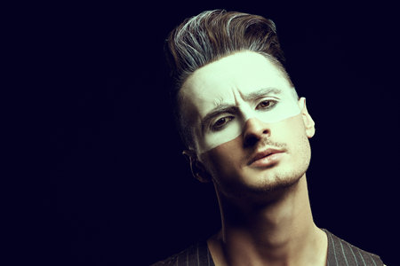 Art portrait of a handsome young man with white paint on his face on a black background with light. Male beauty, fashion. Hairstyle and cosmetics.の写真素材