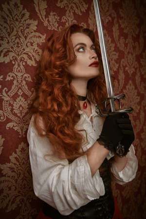 Beautiful and brave red-haired woman looking up with a battle epee (rapier) on a vintage background. The heroine of a historical adventure novel.の写真素材