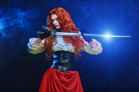 Portrait of a beautiful red-haired girl with a battle epee (rapier) on a blue smoky background. Historical reconstruction of the 16-17th centuries.の写真素材