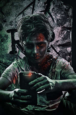 Horrible Halloween. Scary zombie boy teenager boy is on the phone from behind the headstone in the cemetery.の写真素材