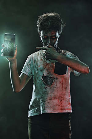 Horrible Halloween. Scary zombie boy teenager boy is on the phone from behind the headstone in the cemetery.の写真素材