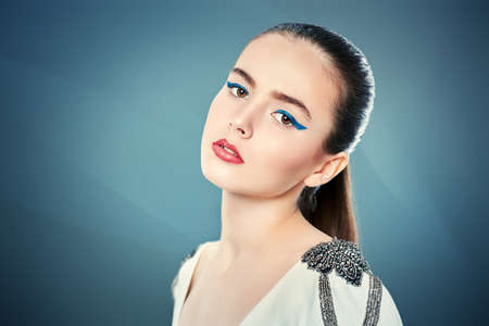 Fashion shot. Portrait of a beautiful young woman in white jacket on a gray background. Trendy makeup with blue aquamarine eye arrows. Make-up and cosmetics.の写真素材