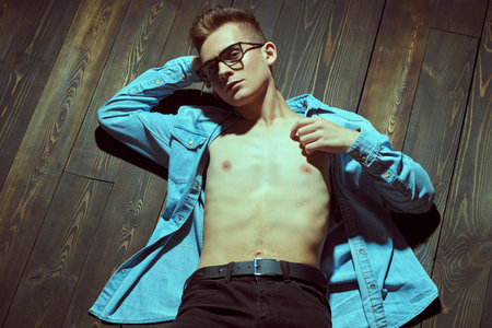 Attractive young man in a denim shirt with a naked torso is lying on the floor. Men's beauty,...