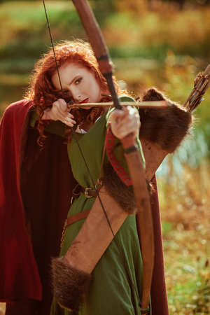 Portrait of a beautiful red-haired girl archer of the Middle Ages stands next to the lake. Celtic culture. Fantasy world.の写真素材