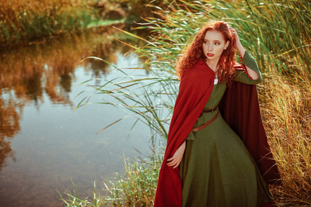 Beautiful red haired girl in a traditional green celtic dress and a red cloak stands by the lake....