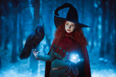Halloween. Pretty young girl witch with a black raven and magic ball in a deep forest. Fairy tales.の写真素材