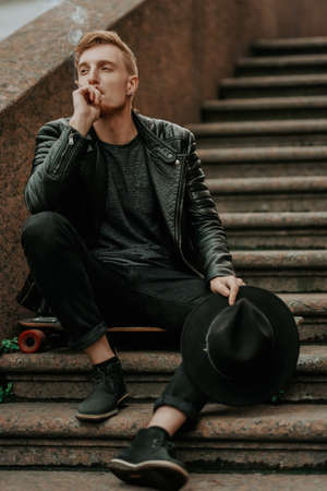 Modern young man hipster sitting on stairs and smoking a cigarette on the street. Full length portrait. Street style.の写真素材