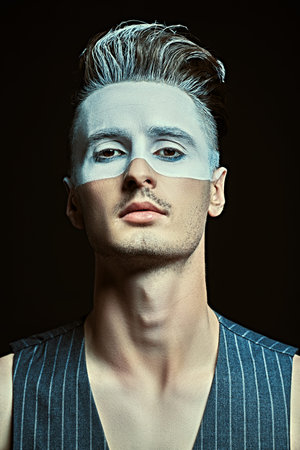 Art portrait of a handsome young man with white paint on his face on a black background with light....