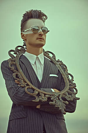 Men's fashion concept. Portrait of a handsome man in elegant suit and trendy hairstyle posing with vintage picture frame against the sky. Body painting.の写真素材