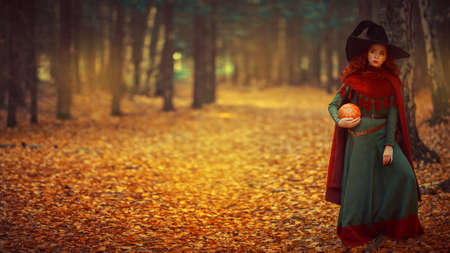 Halloween. Charming young girl witch with long red hair stands in a deep forest with a pumpkin. Fairy tales. Copy space.の写真素材
