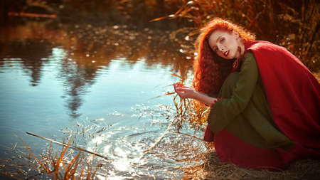 Beautiful red haired girl in a traditional green celtic dress and a red cloak sits by the lake. Historical reconstruction of ancient Celtic times.の写真素材
