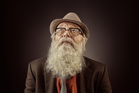 Intelligent old man with long white beard wearing hat and glasses looking up. Studio portrait on a...