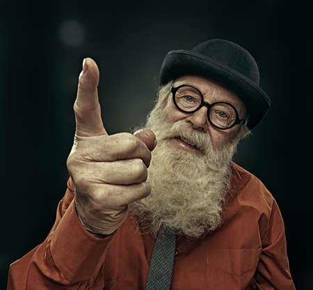 An old man with a long white beard in a bowler hat teaches by threatening with his index finger. Studio portrait on a black background.の写真素材