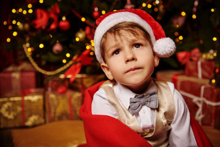Dreamy little boy sits in a Christmas hat under a plaid and looks up. Christmas and New Year concept.の写真素材