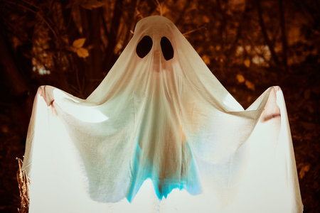 A ghost child under a white sheet with light inside in a dark forest. Halloween concept.