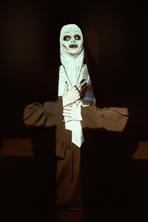Scary devilish nun prays standing in a dark room in light in the form of a cross. Horrors and...