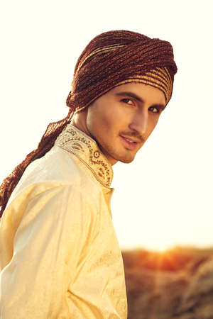 Portrait of a handsome young man in national Indian costume in the desert at sunset. Men's beauty, fashion.の写真素材