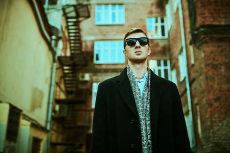 Urban men's style. Handsome man in black sunglasses and elegant black coat stands against the background of the grunge brick houses of the big city.の写真素材