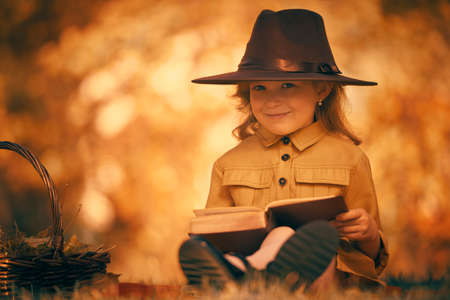 Pretty little girl is reading a book sitting on a blanket on the grass next to a basket of leaves. Retro style. Children's fashion.の写真素材