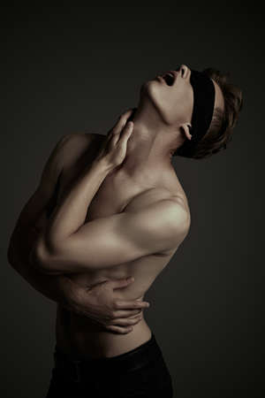Art portrait of a handsome blindfold young man with perfect muscular body raised his head up, expressing tension and suffering. Black background, darkness.の写真素材