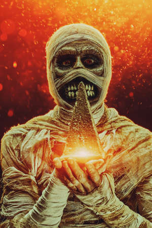 Scary evil mummy is holding small copy of pyramide in his hands in a magical sparkling light. Halloween. Ancient Egyptian mythology.の写真素材