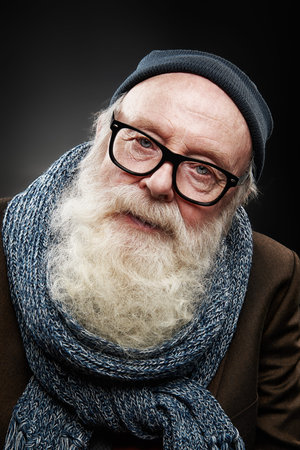 An old man with a long white beard in a hat and a scarf looks at the camera carefully. Studio...