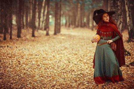 Halloween. Charming young girl witch with long red hair stands in a deep forest with a pumpkin....