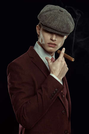 Handsome young man in an expensive three-piece suit and a cap smoking a cigar on a black background. Retro style, mafia man. Men's beauty, fashion.の写真素材