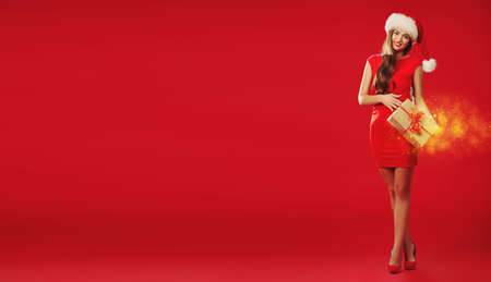 Gifts on Christmas and New Year. Full length portrait of a beautiful smiling girl in red dress and Santa Claus hat holding a gift box. Studio shot over festive red background. Copy space.の写真素材
