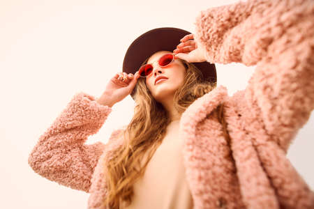 Autumn fashion. Stylish young woman in a fashionable fur coat and hat looks at the sky through pink glasses.の写真素材
