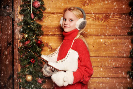 Winter activities. Happy girl child in winter clothes is going to skate. She stands under snowfall by a wooden house decorated with Christmas spruce garlands and toys.の写真素材