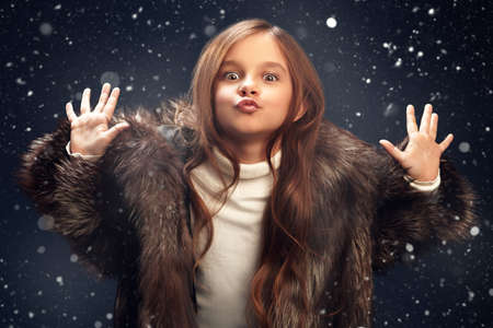 Funny emotional little girl in a silver fox fur coat stands under the snowfall. Christmas winter kids fashion.の写真素材