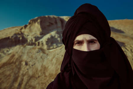 Courageous young man in black clothes and black keffiyeh on his head stands in a desert and looks pensive forward. Adventures, the spirit of freedom.の写真素材