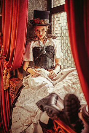 Portrait of a charming steampunk lady sitting on a windowsill in a room with Victorian vintage...