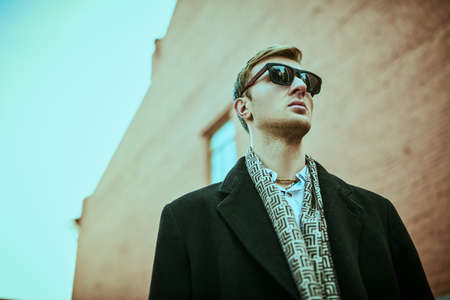 Urban men's style. Handsome man in black sunglasses and elegant black coat stands against the background of the grunge brick houses of the big city.の写真素材