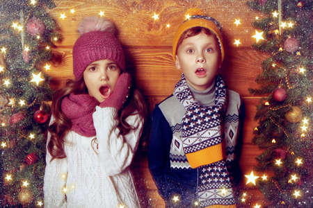 Two cute kids in winter clothes stand by a wooden wall beautifully decorated for Christmas and are surprised. Snowflakes and shining stars are around.の写真素材