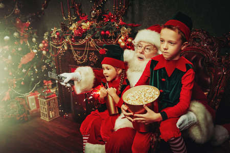 Cheerful Santa Claus and elves watching a movie with soda and popcorn in a house beautifully decorated for Christmas. Entertainment and cinema concept.の写真素材