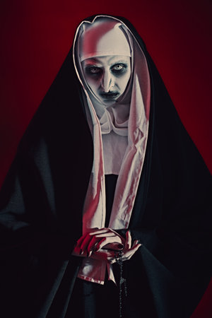 Scary devilish possessed nun with a cross in her hands standing on a bloody red background. Horrors and Halloween.の写真素材