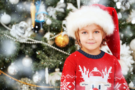 Portrait of a cute happy little boy in Santa Claus hat smiling at camera. Beautiful Christmas tree...