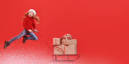 Happy girl child in Christmas hat pushing the sled with gifts. Studio portrait over festive red...