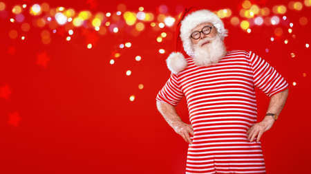 Portrait of Santa Claus in old-fashioned striped sleepwear stands on a red background with festive lights. Copy space. Christmas and New Year.の写真素材