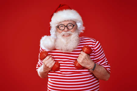 Funny Santa Claus doing exercises with dumbbells on a festive red background. Sport and healthy lifestyle. Copy space.の写真素材