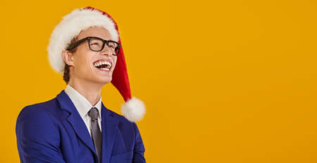 Christmas and New Year celebration. Happy young man in elegant formal suit and Santa Claus hat is laughing. Yellow background with copy space.の写真素材