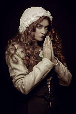 Sepia portrait of a praying woman with long red hair wearing medieval clothes on a black background. Historical reconstruction of the Middle Ages.の写真素材
