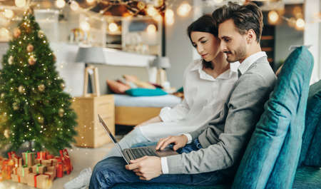 Young couple in love resting at home for Christmas. They use a laptop while sitting on the couch in a festive atmosphere.の写真素材
