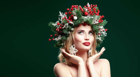 Portrait of a charming blonde woman with christmas wreath on her head and snowflake earrings dreamily looking up and smiling. Green background with copy space.の写真素材