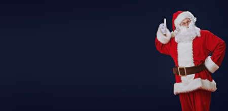 Attention, Christmas! Santa Claus raises his index finger to attract attention. Studio portrait over dark blue background. Copy space.の写真素材